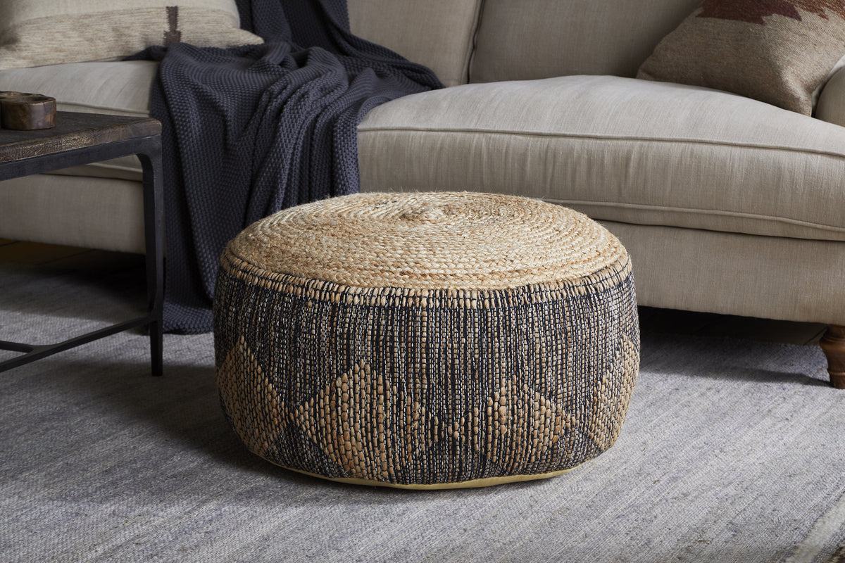 The Kapil Jute Round Pouf - Charcoal by nkuku, featuring a geometric braided design, sits on a light gray rug in front of a beige sofa with a dark blanket, while a wooden coffee table adds to the chic setting.