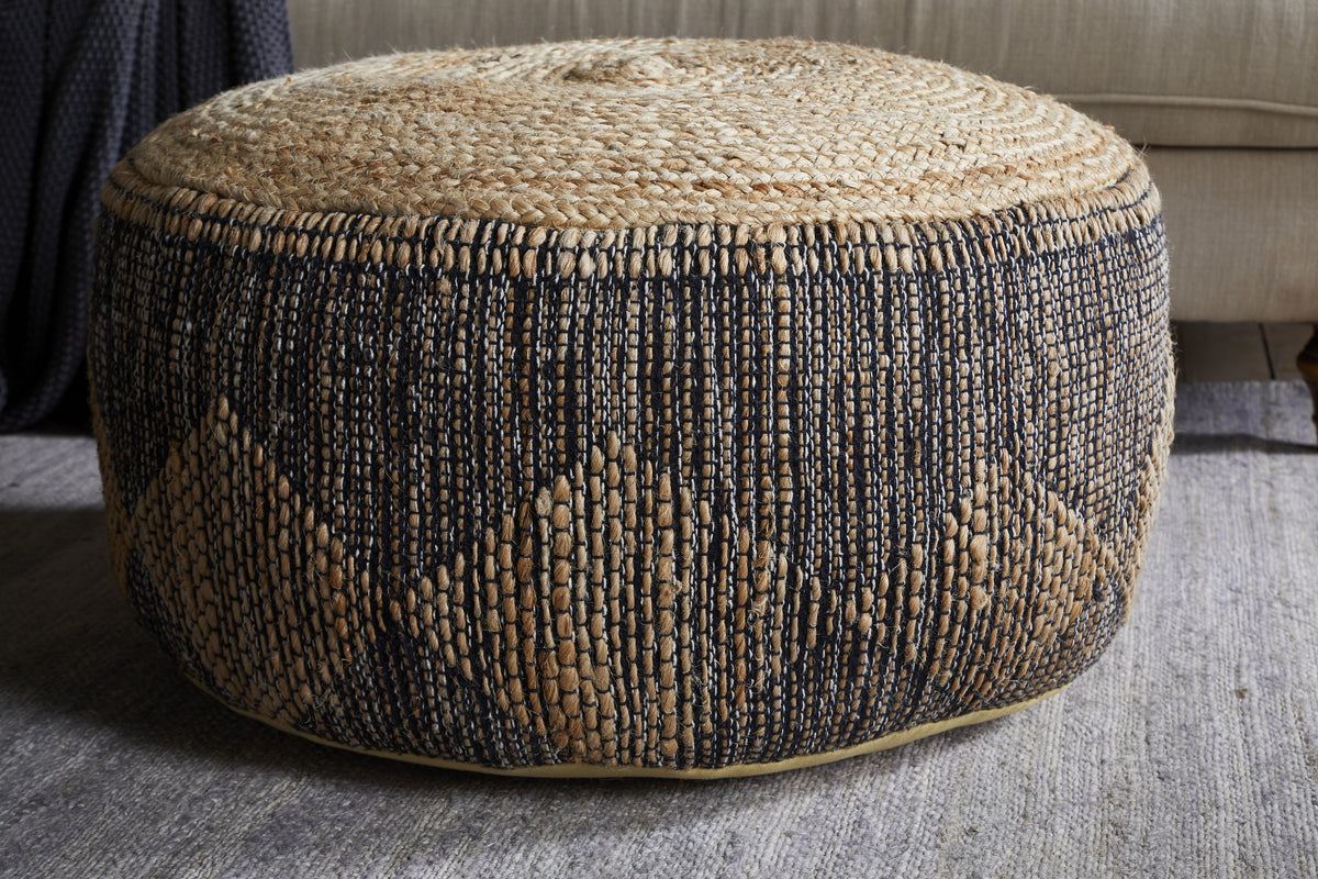 The Kapil Jute Round Pouf - Charcoal by nkuku, featuring a natural tan top and black diamond braided design, sits on a gray rug in front of a beige sofa, adding cozy texture and style to your living space.