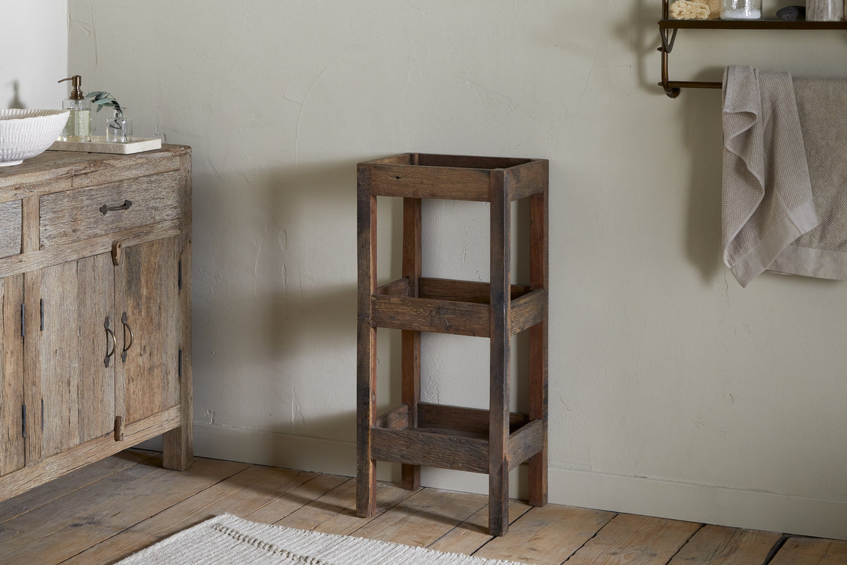 The Akara Reclaimed Wood Storage Caddy by nkuku sits in a bathroom corner with light walls. To the left is a wooden vanity with a white sink and glass soap dispenser, while a towel hangs on a metal rack above wooden flooring.