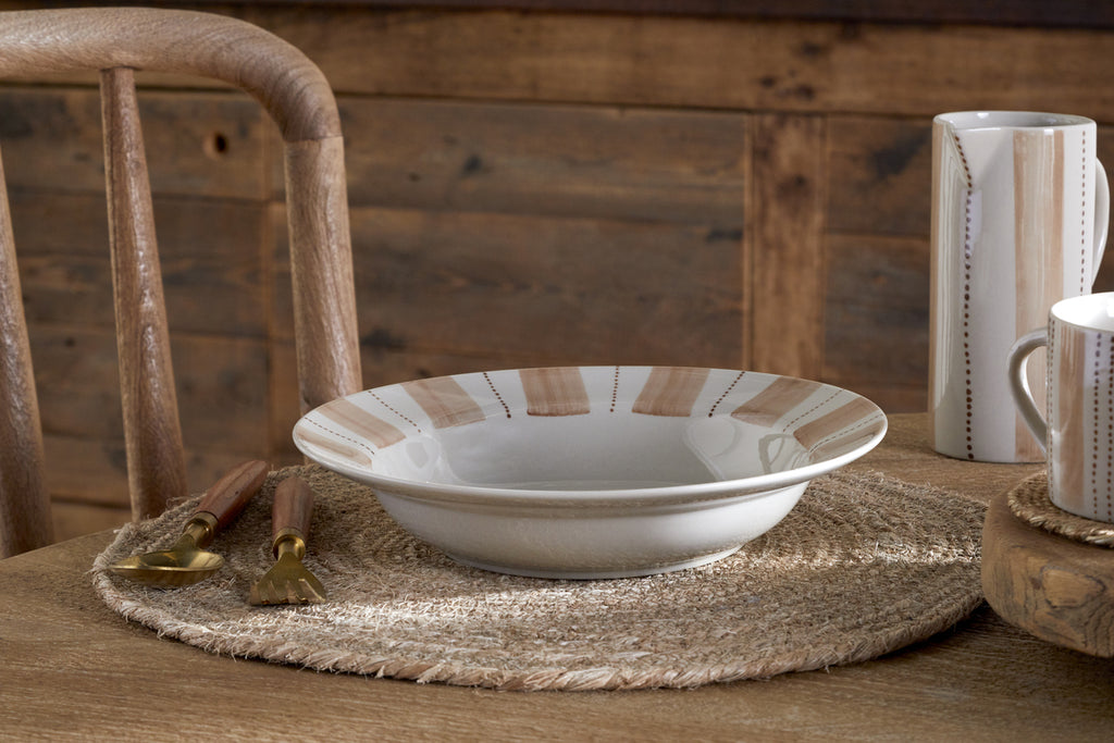 The nkuku Patthar Pasta Bowl - Cream & Clay, with beige and tan stripes, rests on a wooden table with a fork and spoon, plus a matching striped cup and pitcher from the same collection.