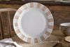 The nkuku Patthar Dinnerware Set (Cream & Clay, Set of 12) features round white ceramic plates with tan and beige borders, displayed on a woven placemat with matching hand-painted bowls stacked nearby. A wooden chair and rustic table complete the scene.