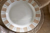 The Patthar Dinner Plate - Cream & Clay by nkuku, featuring a hand-painted rim of beige rectangles and dotted lines on white ceramic, sits empty on a woven placemat atop a wooden table beside striped cloth and bread.
