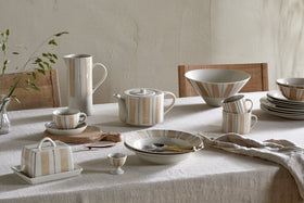 A table set with nkuku's Patthar Dinner Plate - Cream & Clay, featuring hand-painted beige and brown stripes. The cream ceramic set includes teapot, cups, bowls, and plates on a beige tablecloth, plus a vase with green sprigs in the background.