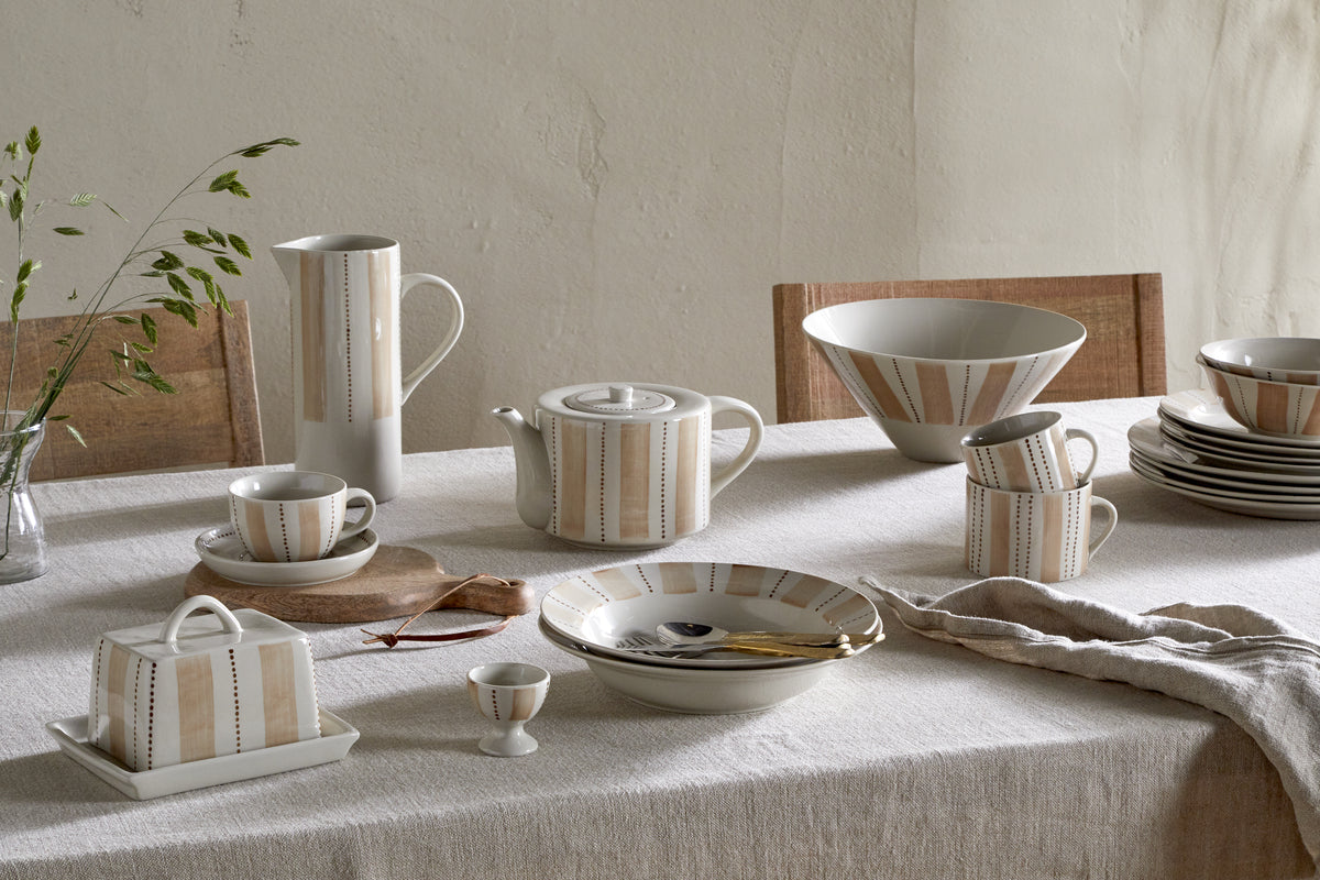 A table set with nkuku's Patthar Dinner Plate - Cream & Clay, featuring hand-painted beige and brown stripes. The cream ceramic set includes teapot, cups, bowls, and plates on a beige tablecloth, plus a vase with green sprigs in the background.