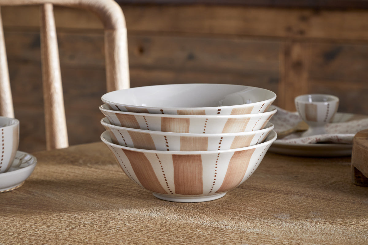The Patthar Dinnerware Set - Cream & Clay by nkuku features 12 hand-painted ceramic pieces, stacked on a wooden table with rustic surfaces in the background, evoking a cozy farmhouse kitchen vibe.