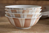 Four nkuku Patthar Cereal Bowls in Cream & Clay are stacked on wood. Each hand-painted bowl features vertical beige stripes and dotted lines, with the warm, blurred wooden background highlighting the artisan dinnerware’s charm.