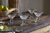 A set of four nkuku Sooraj Champagne Coupes (Clear) sits on a rustic wooden table, surrounded by lime wedges, a small bowl of salt, and fresh limes and lemons in the background for a vintage-inspired display.