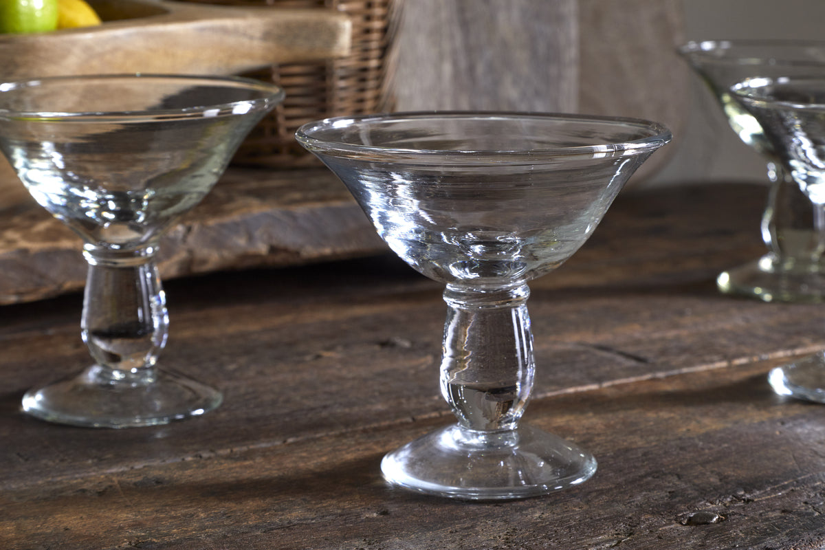 The nkuku Sooraj Champagne Coupe set of 4 in clear glass features wide, shallow bowls and short stems. Displayed on a rustic wooden surface with fruit and a woven basket, these coupes exude timeless vintage charm.