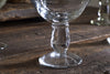 Close-up of the clear stem and base of an nkuku Sooraj Champagne Coupe (Set of 4), resting on a rustic wooden table with a softly blurred background that highlights the glass and wood textures.