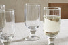 Four Sooraj Large Glass goblets by nkuku, from a set of 4, rest on a light tablecloth. Three are empty; the rightmost is half-filled with a pale summer drink. A simple, neutral background hints at a wooden chair.