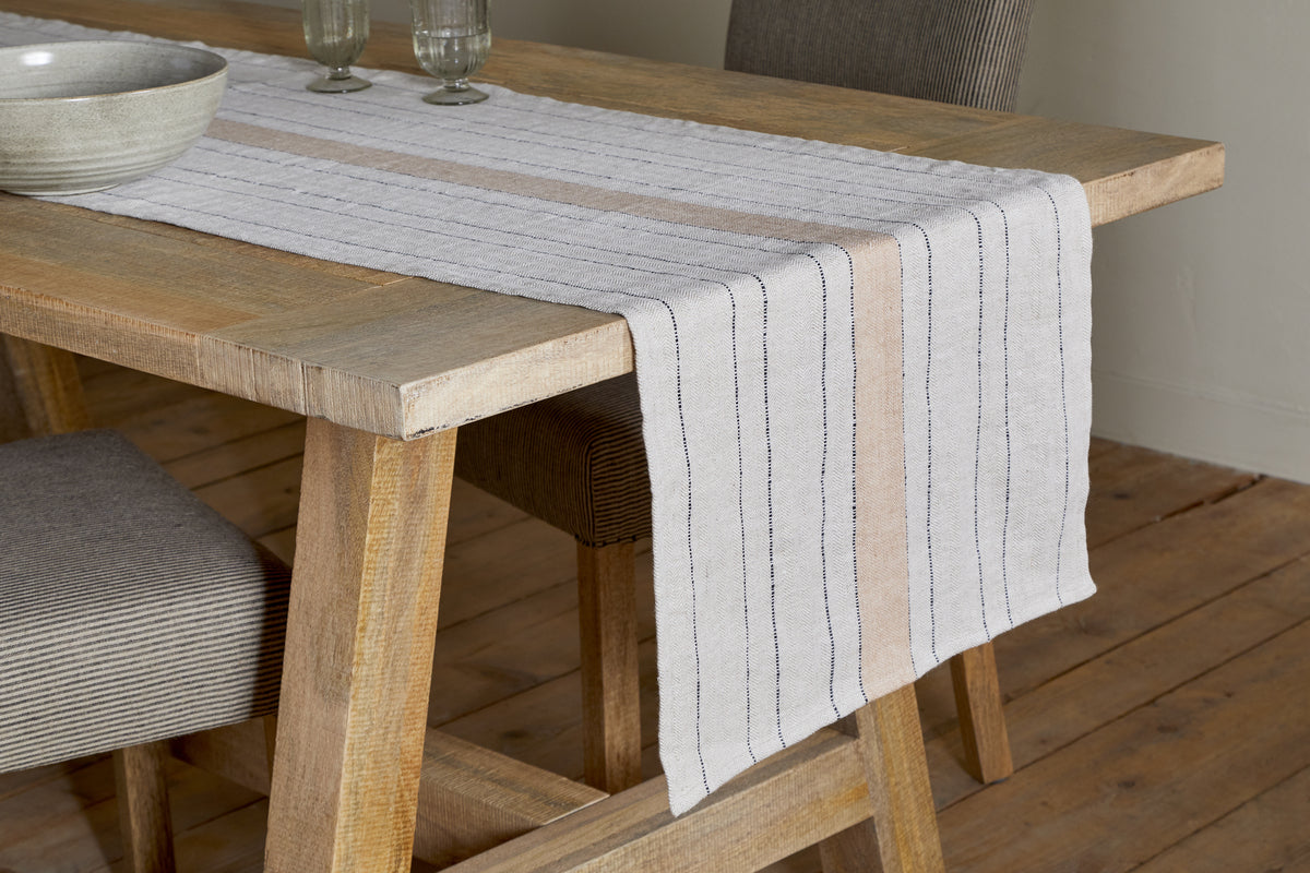 A rustic wooden dining table features the Buna Table Runner - Clay by nkuku with black and tan stripes, two glass goblets, and a large light-gray ceramic bowl, paired with striped chairs on a light wood floor.