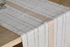A close-up of the nkuku Buna Table Runner - Clay, featuring natural stripes—thin black and one wide beige—draped over a wooden dining table, with a glass partially visible in the top left corner.