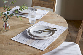 A round wooden table set for two features nkuku Buna Placemats in Clay, stacked white plates, cutlery, water glasses, a small vase with green leafy branches, and is surrounded by upholstered wooden chairs.