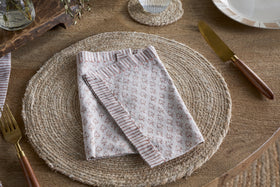A beige woven placemat sits on a wooden table with an nkuku Peplu Block Print Napkin - Terracotta folded at the center. A gold fork is on the left, a gold knife with a wooden handle on the right, and part of a white plate is visible in the top right corner.