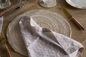 A rustic table setting with a round woven placemat, nkuku Peplu Block Print Napkin in Terracotta, gold cutlery with wooden handles, and a clear glass on a matching coaster atop a wooden tabletop.