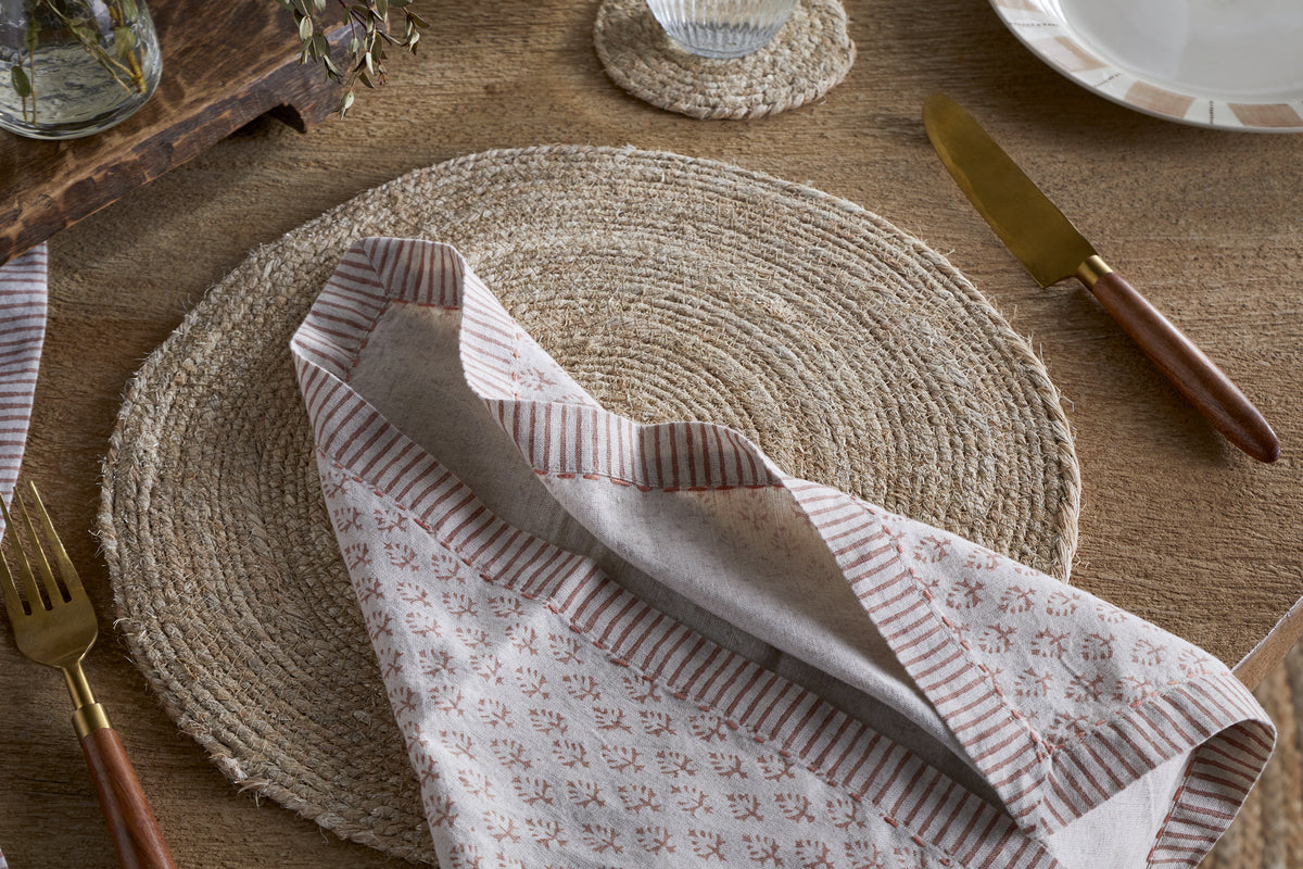 A rustic table setting with a round woven placemat, nkuku Peplu Block Print Napkin in Terracotta, gold cutlery with wooden handles, and a clear glass on a matching coaster atop a wooden tabletop.