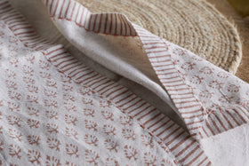 Close-up of nkuku's Peplu Block Print Napkin in Terracotta (Set of 4), showcasing brown floral and stripe patterns on beige fabric with stitched seams, resting on a woven placemat in soft natural light for a rustic look.
