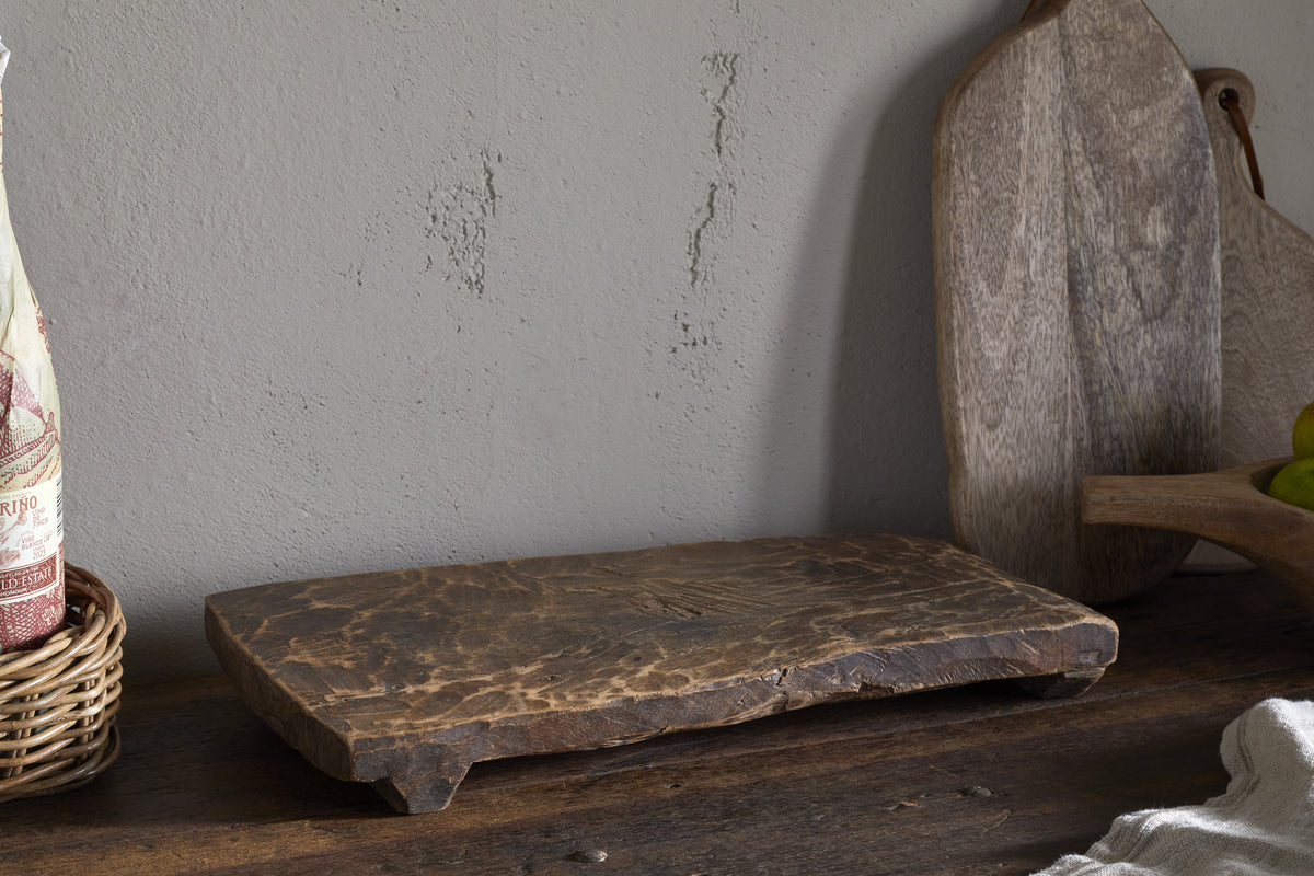 The Kaveeta Reclaimed Serving Board - Natural by nkuku, with its rustic, textured wood, rests on a dark table. Behind it, two lighter wood boards lean against a gray wall, with hints of a wicker basket and linen cloth nearby.