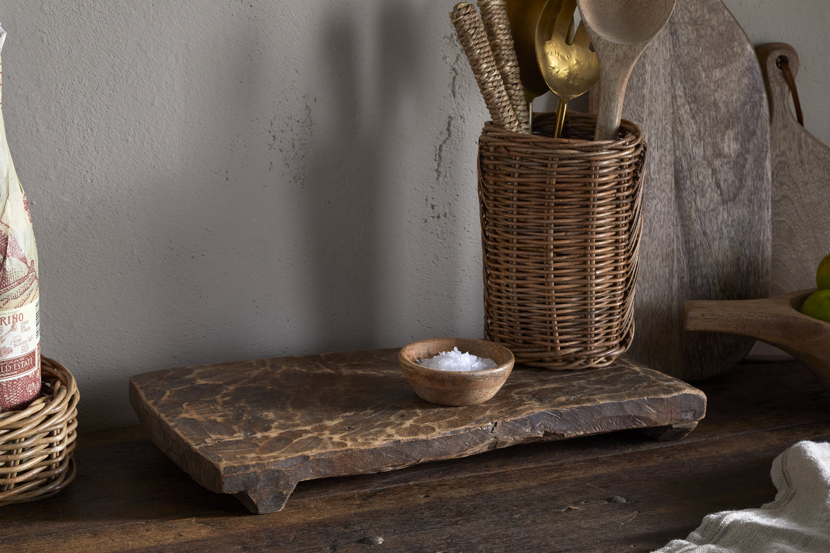 A rustic kitchen scene with a small wooden bowl of salt on the Kaveeta Reclaimed Serving Board - Natural by nkuku. Behind it, a wicker utensil holder with wooden and brass utensils stands beside a large round cutting board against a light grey wall.