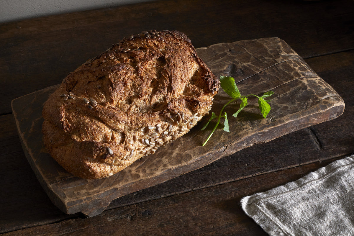 A rustic seeded brown bread sits on the Kaveeta Reclaimed Serving Board - Natural by nkuku, with a sprig of fresh herbs beside it. The scene features a dark wooden table and a folded beige cloth in the corner.
