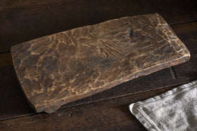 The Kaveeta Reclaimed Serving Board - Natural by nkuku, featuring a rustic rectangular design with visible grain and character markings, rests on a dark wood surface beside a light beige textured cloth for vintage appeal.