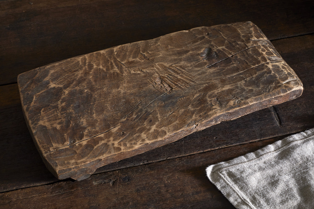 The Kaveeta Reclaimed Serving Board - Natural by nkuku, featuring a rustic rectangular design with visible grain and character markings, rests on a dark wood surface beside a light beige textured cloth for vintage appeal.