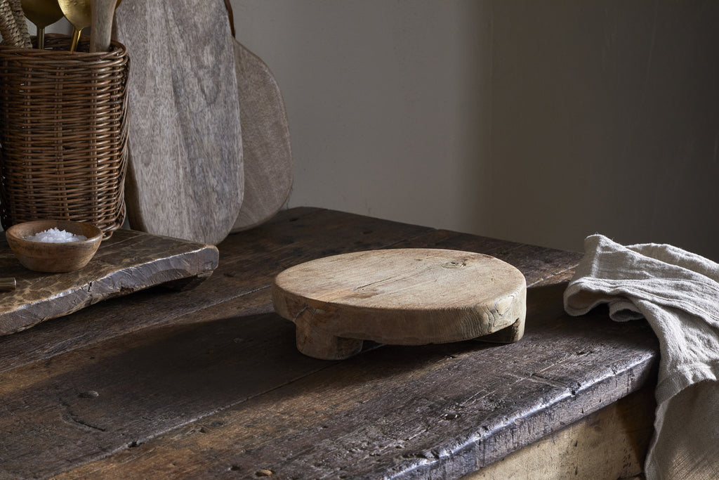 The nkuku Kaveeta Board - Natural, crafted from reclaimed wood with short legs, rests on a rustic wooden table, surrounded by a beige cloth and utensils basket. A small bowl of coarse salt sits on the board, softly lit for warmth and patina.