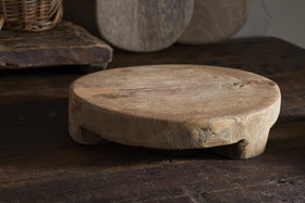 The Kaveeta Board - Natural by nkuku features a round, rustic wood design with visible grain and timeworn details, resting on a dark surface. Reclaimed boards and baskets in the background create an earthy, vintage feel.