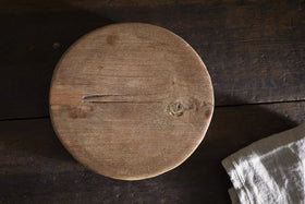 The Kaveeta Board - Natural by nkuku, a round reclaimed wood board with rustic grain, cracks, and knots, sits on a dark wood surface. A light beige textured cloth is visible in the image's bottom right corner.