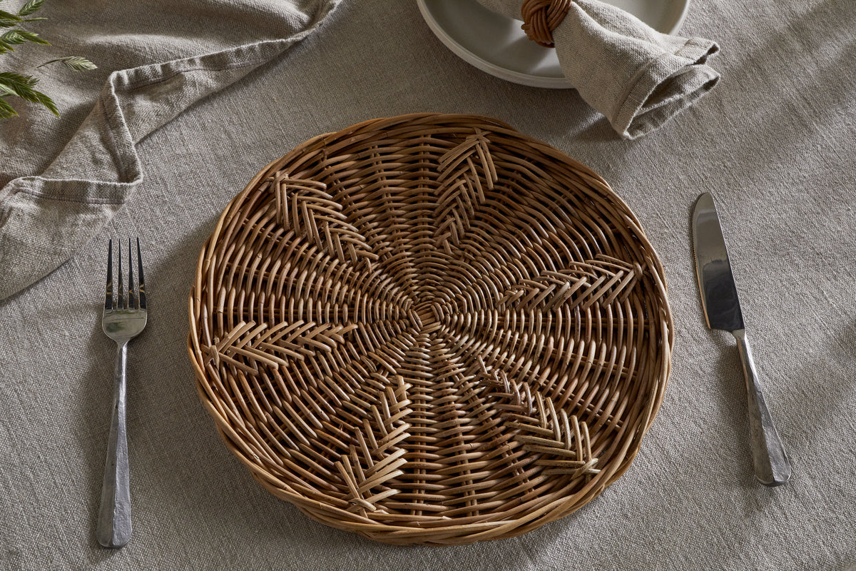 The nkuku Bersama Rattan Placemat - Natural sits on a beige linen tablecloth, with cutlery on each side. A beige napkin in a rattan ring tops stacked plates, creating a rustic dining setting.