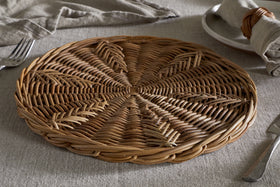 The Bersama Rattan Placemat by nkuku, featuring intricate handwoven details, rests on a light gray tablecloth beside a plate with a napkin in a wicker ring, fork, and knife—creating a rustic dining setting in soft natural light.