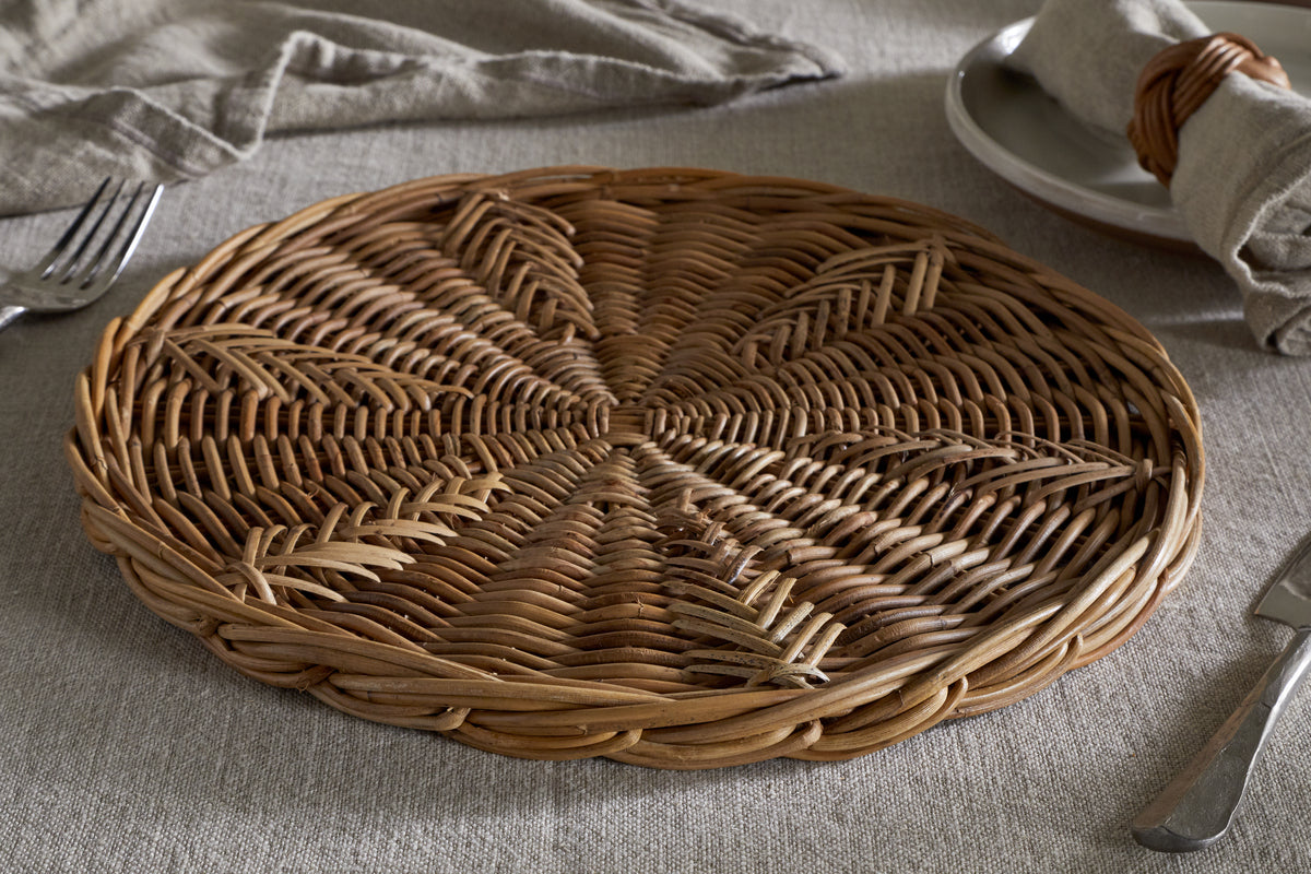 The Bersama Rattan Placemat by nkuku, featuring intricate handwoven details, rests on a light gray tablecloth beside a plate with a napkin in a wicker ring, fork, and knife—creating a rustic dining setting in soft natural light.