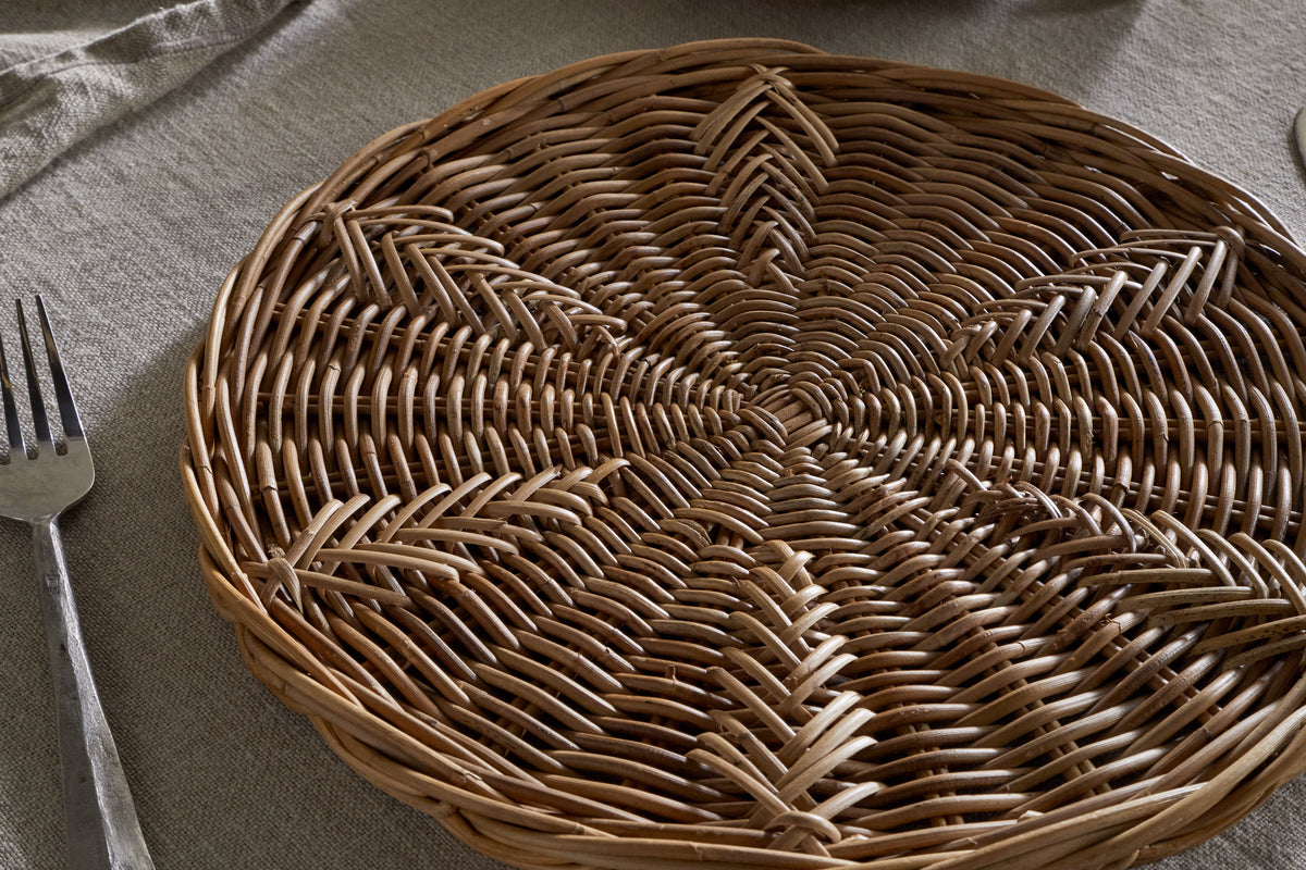 The nkuku Bersama Rattan Placemat - Natural, featuring intricate handwoven details, rests on a beige fabric tablecloth. A metal fork lies to the left, with a folded napkin partly visible in the upper left corner of the table.