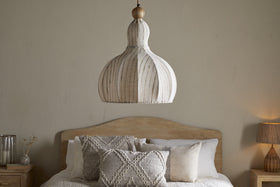 A neutral-toned bedroom features a wooden bed with cream and gray pillows. The nkuku Kela Fabric Pendant in Off White hangs above, while a lamp and glass vase rest on the light wood nightstand.