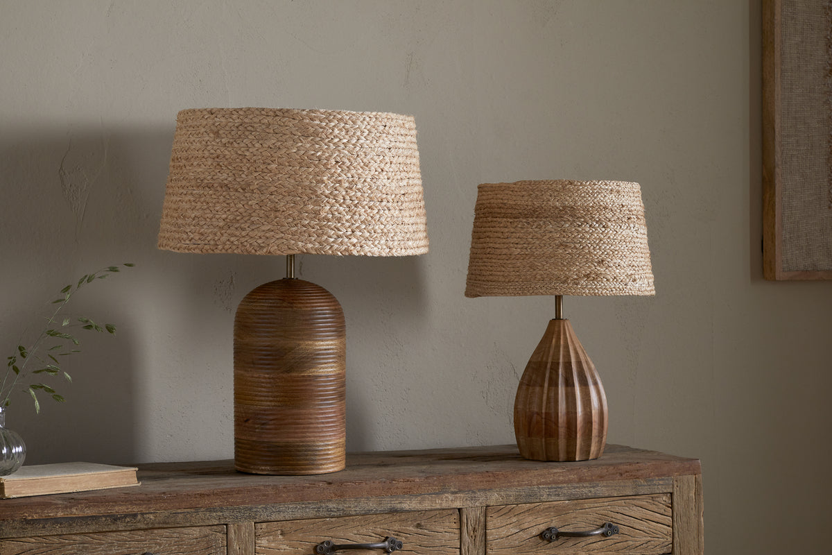 Two table lamps with nkuku Pisang Natural Lampshades sit on a rustic dresser. The left lamp has a rounded, ridged base; the right is smaller with a striped, vase-shaped base. A sprig in a glass and a closed book rest nearby.