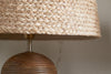 Close-up of the nkuku Pisang Natural Lampshade - Natural, featuring a woven jute shade and ribbed wooden base, set against a light-colored wall with a visible crack.