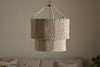 The Thimba Woven Jute Chandelier by nkuku hangs from the ceiling against a plain, light wall. Below, a neutral sofa with textured pillows adds rustic coziness and minimalist charm to the room.