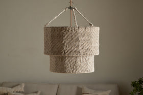 The Thimba Woven Jute Chandelier by nkuku hangs from the ceiling against a plain, light wall. Below, a neutral sofa with textured pillows adds rustic coziness and minimalist charm to the room.