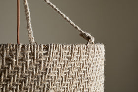 Close-up of the Thimba Woven Jute Chandelier by nkuku, showcasing its rustic texture, zig-zag pattern, and thick rope handle threaded through a metal ring. Set against a neutral beige background with gentle natural light.