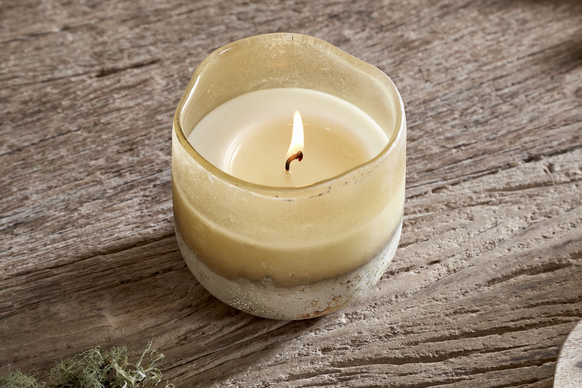 The nkuku Oakmoss & Rose 1 Wick Scented Candle - Yellow sits in a rustic, yellow glass holder with an uneven rim and frosted, textured base. Its steady flame casts a warm glow on the weathered wood beneath.