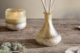A candle and the nkuku Oakmoss & Rose Scented Reed Diffuser - Yellow fill your space with soft light and a floral woody scent, creating a cozy ambiance.