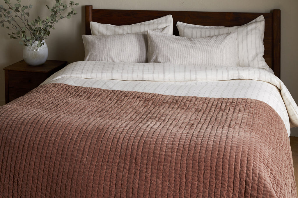 A neatly made bed with white and striped pillows, a white and beige duvet, and the nkuku Nidra Velvet & Linen Bed Quilt – Rust featuring Kantha quilting, next to a wooden nightstand with eucalyptus—a perfect King Super King bedding set.