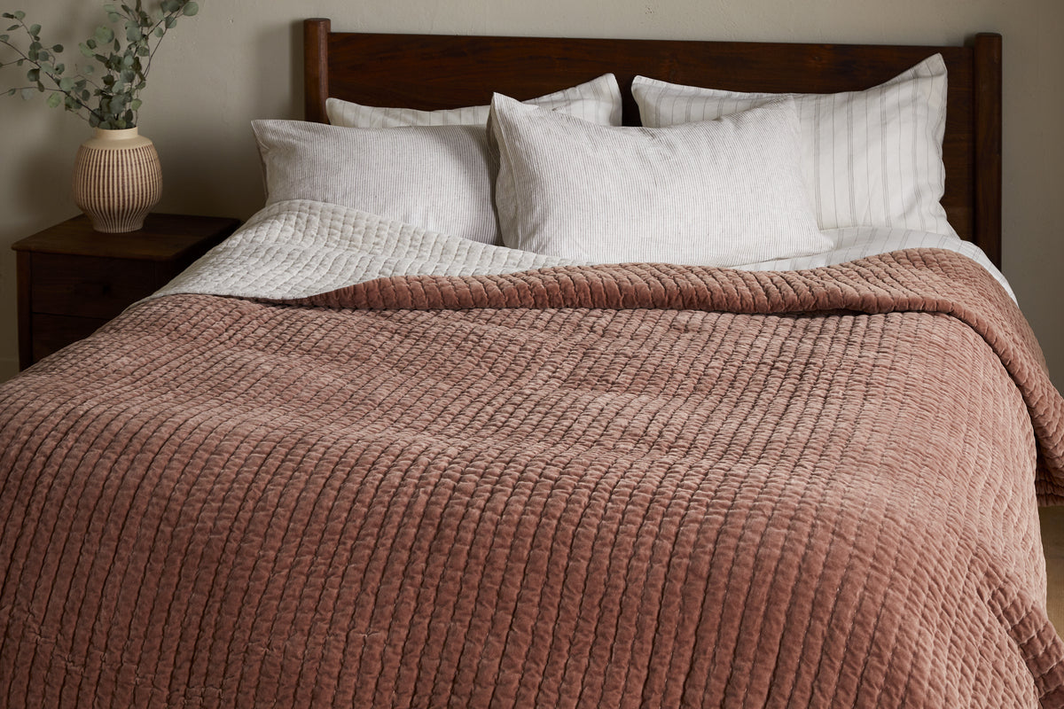A neatly made bed with white pillows, a striped white sheet, and the nkuku Nidra Velvet & Linen Bed Quilt in Rust. A wooden headboard and a nightstand with greenery complete this King Super King bedding setup.