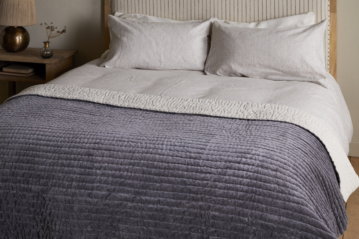 The nkuku Nidra Velvet & Linen Bed Quilt in Charcoal, with Kantha quilting, is styled on a neatly made bed with white pillows and a beige blanket. A wooden nightstand with a vase and lamp sits beside—ideal for King and Super King beds.