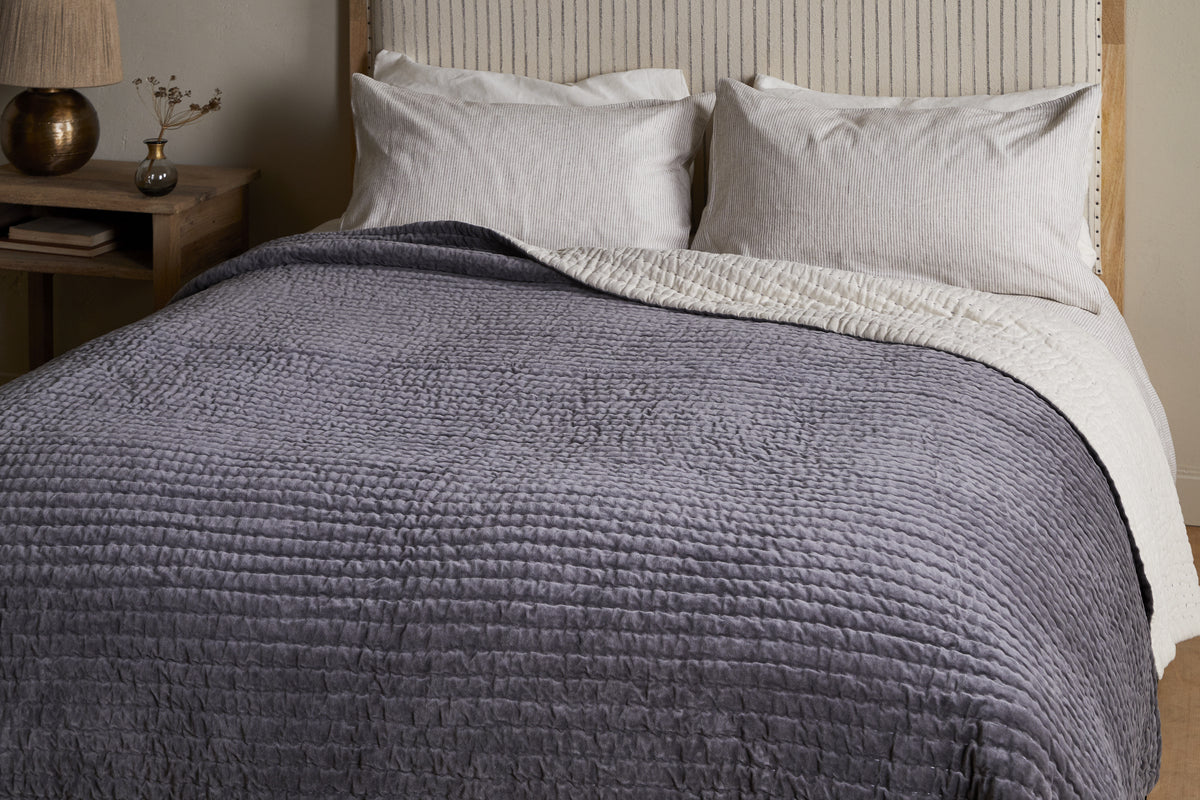 A neatly made bed features the nkuku Nidra Velvet & Linen Bed Quilt in Charcoal, layered over a white blanket with two large white pillows and a beige headboard. Ideal for King and Super King beds, with a wooden nightstand, vase, and lamp beside it.