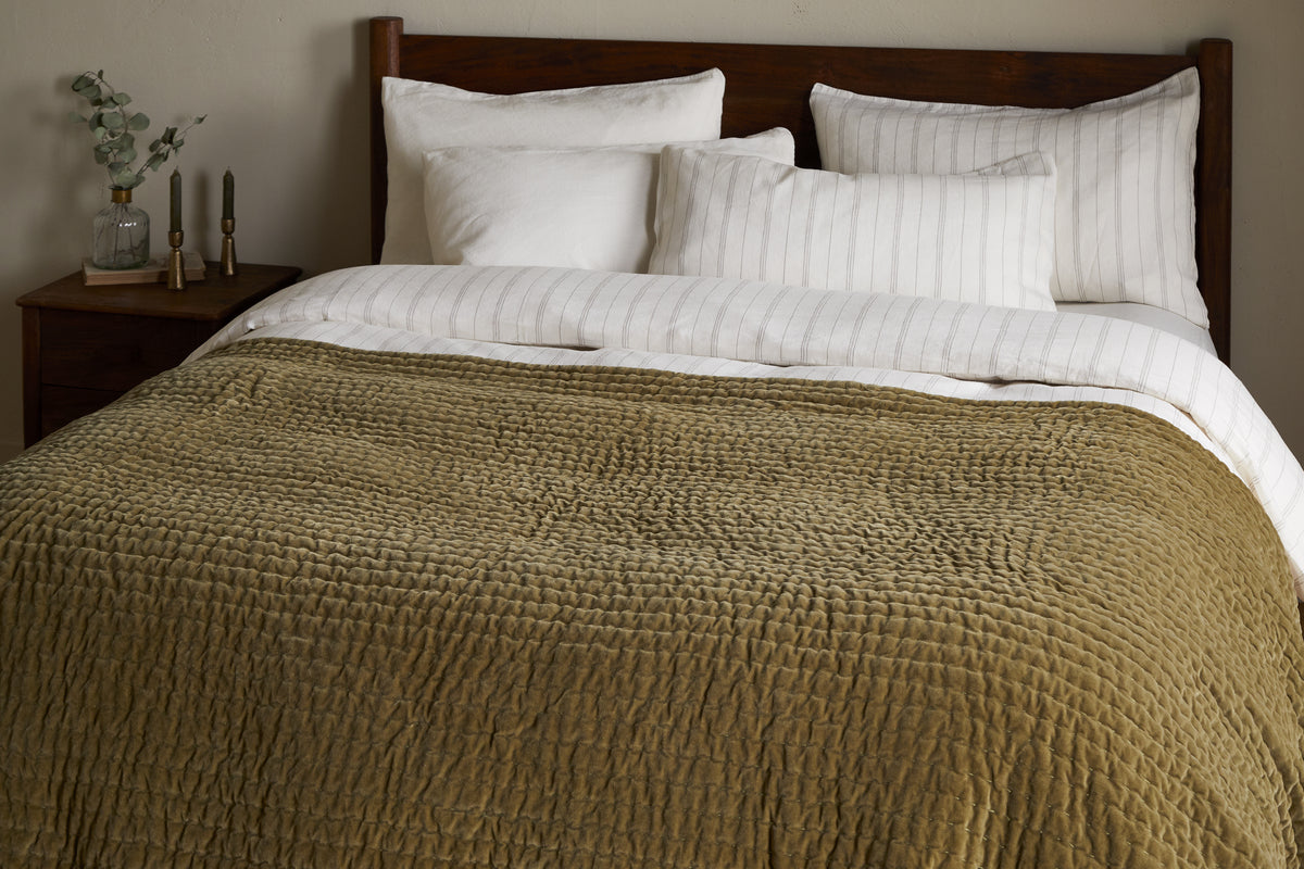 A king size bed is dressed with white pillows, a cream-striped blanket, and the nkuku Nidra Velvet & Linen Bed Quilt in Moss, featuring Kantha quilting. A wooden headboard and nightstand with decor items are in the background.