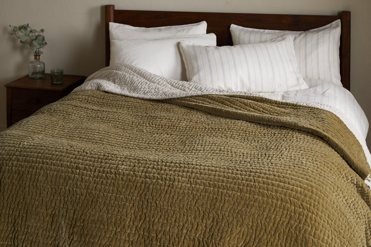 A neatly made bed with white pillows and linens is partially covered by the nkuku Nidra Velvet & Linen Bed Quilt in Moss. Against a wooden headboard, a small nightstand holds a glass vase of eucalyptus and a candle.