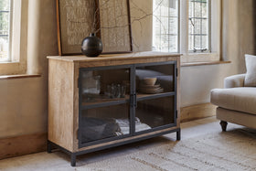 The nkuku Nirvi Mango Wood & Iron Sideboard - Black features glass doors displaying glassware and plates. A black vase with branches and framed art sit atop, next to a window and beige sofa on a woven rug in a sunlit room.