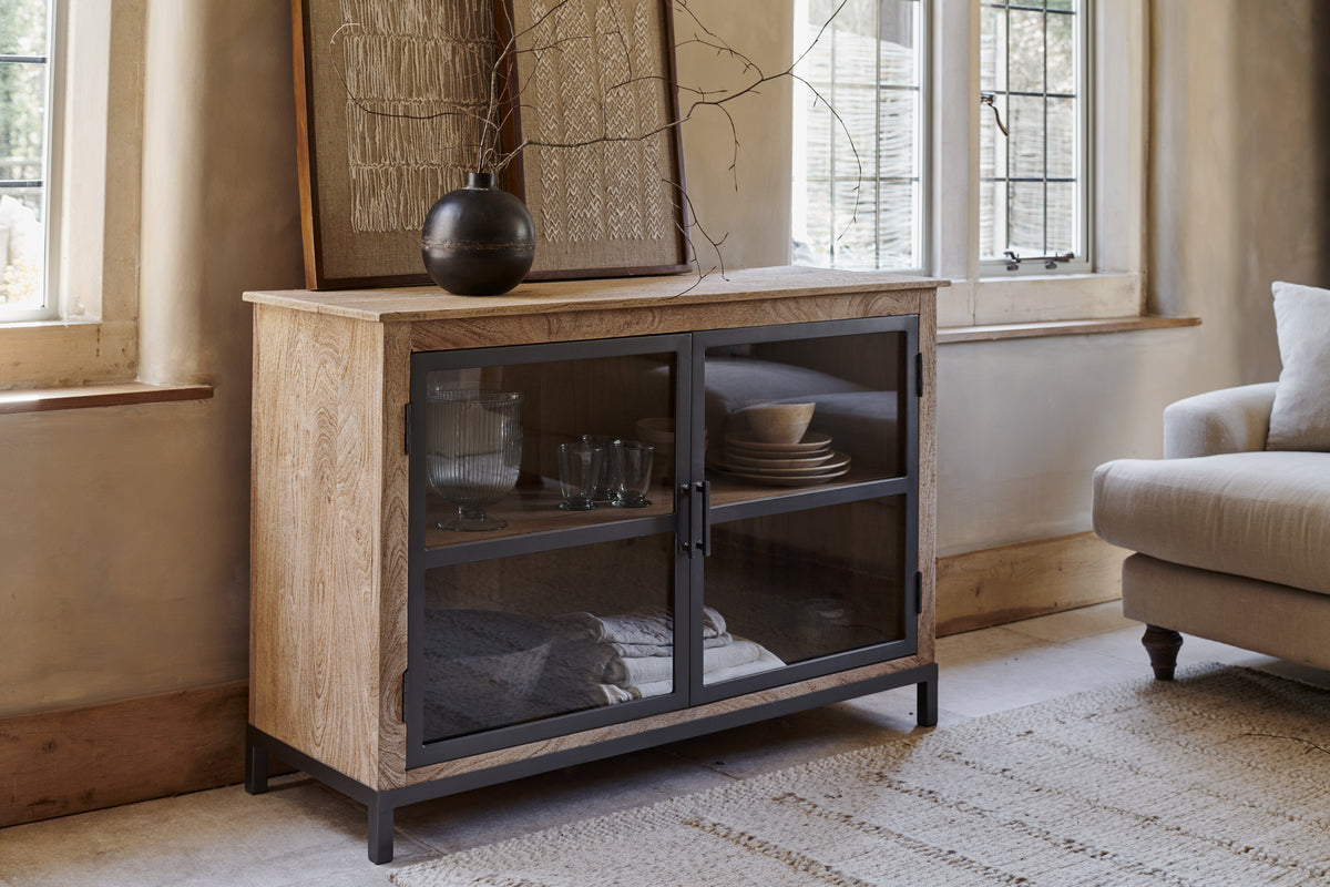 The nkuku Nirvi Mango Wood & Iron Sideboard - Black features glass doors displaying glassware and plates. A black vase with branches and framed art sit atop, next to a window and beige sofa on a woven rug in a sunlit room.
