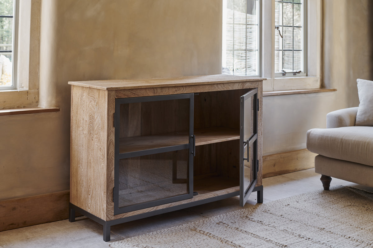 The Nirvi Mango Wood & Iron Sideboard - Black by nkuku, featuring black metal-framed glass doors, stands beside a window in a bright room. One door is open to show a shelf inside; a light armchair and rug are partly visible.